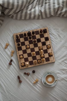 Chess board with cup of coffee on bed. Strategy thinking. Morning ideas concept. Pastel shades. High quality photo. Top view of unmade bedding sheets and pillow.