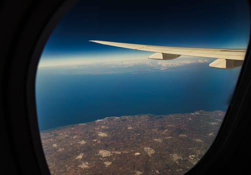 Beautiful view of the blue sea and part of the land with a wing through the airplane window in the evening summer time, close-up side view.