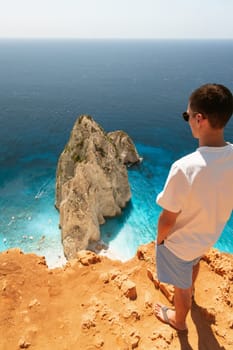 One Caucasian girl in light clothes stands half-turned on the edge of the mountain and looks at a rock in the blue sea on a sunny summer day, close-up view from above.