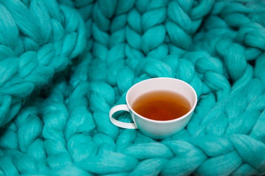 The concept of coziness and comfort is a green knitted blanket on which there is a white cup of tea. Close-up