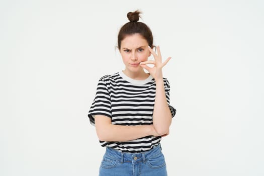 Young woman with doubtful face, zips her lips, seals mouth, promise not to tell anyone, isolated over white background. Copy space