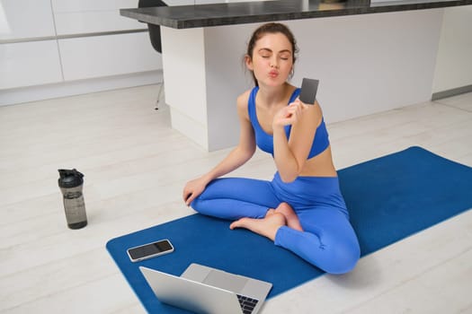 Image of smiling young woman, holding credit card, sitting on fitness mat with laptop, buying sport app subscription, workout video tutorials, doing exercises at home.