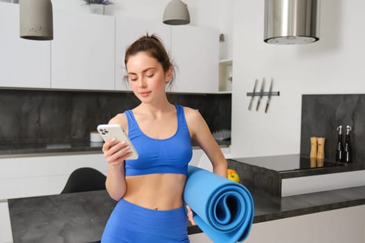 Portrait of attractive, fit young woman in sportswear, standing in living room with rubber yoga mat, holding smartphone, doing fitness exercises with mobile app at home.