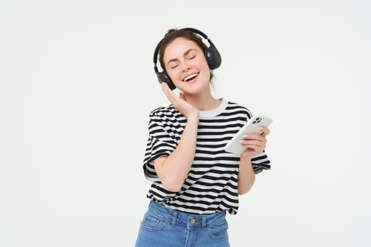 Young woman with smartphone listening to music, dancing to her favourite song in headphones, posing against white background.