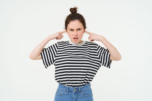 Image of annoyed, bothered woman plugs, shuts her ears with fingers, disturbed by loud noise, stands over white background.