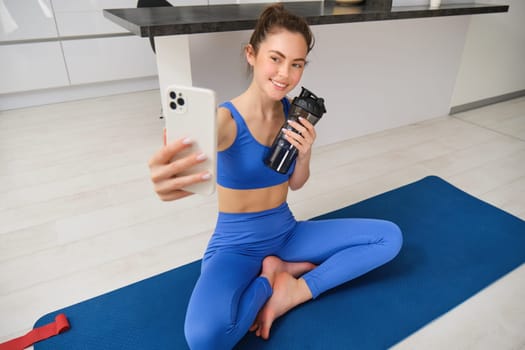 Portrait of woman taking selfie with water bottle, fitness instructor shows her exercises, doing workout from home, on rubber blue yoga mat, wearing sportswear.
