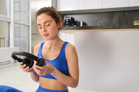 Image of young fitness girl, wearing blue tracksuit, puts on wireless headphones to workout with music, does yoga at home.