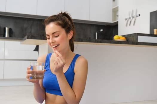 Portrait of healthy, smiling young woman taking vitamins after workout, holding glass of water and dietary supplement, buds for health and energy.