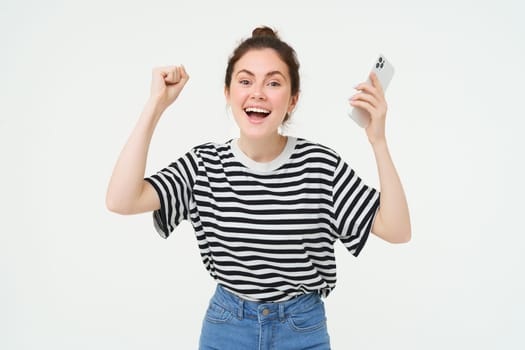 Portrait of happy woman laughing, holding telephone, using smartphone and chanting, rooting for someone, holding hands up, isolated over white background.