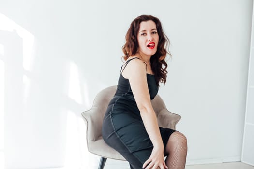 brunette woman in a black dress sits on a chair in a bright room