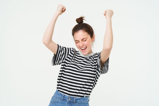 Portrait of happy young sports girl, fan rooting for team, celebrating victory, raising hands up, chanting, triumphing, standing over white background. Copy space