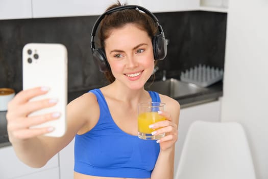 Close up portrait of smiling brunette woman, fitness girl takes selfie for her social media, wears wireless headphones, holds glass of orange juice, stands in kitchen in sportswear.