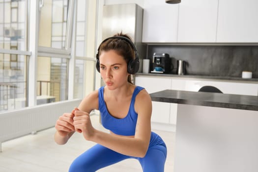 Image of young sportswoman, fitness instuctor doing squats workout at home, listening music in wireless headphones and stretching arms, doing exercises in living room.