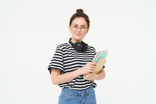 Young woman with notebooks, books and study material, posing over white background, wears headphones over neck.