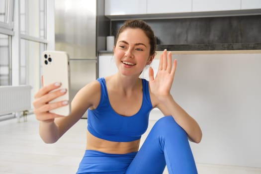 Image of young woman doing workout, takes selfie, records online video. Fitness instructor records her training on phone, says hello at smartphone camera and waves hand at it.