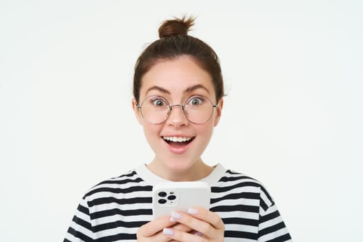 Image of young woman in glasses, using her mobile phone, standing with smartphone and smiling, standing over white background.