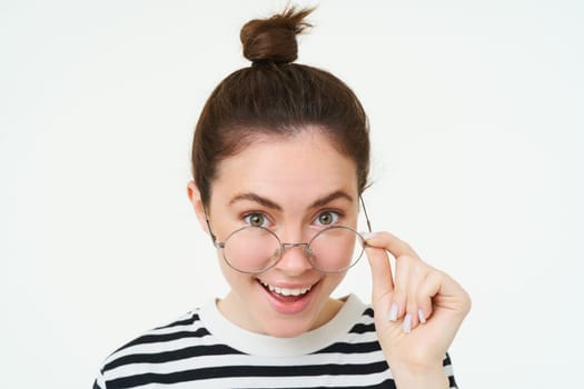 Image of woman in glasses, looks surprised, takes off eyewear, looks amazed, says wow, impressed by something, stands over white background.