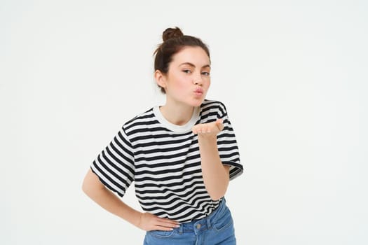 Portrait of cute young woman blows air kiss, sends mwah at camera, posing cute against white background. Lifestyle and people concept.