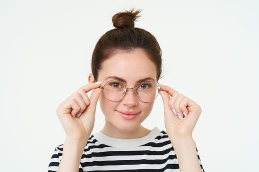 Close up portrait of smiling, attractive young woman wearing new eyewear, trying on glasses for vision, standing over white background.