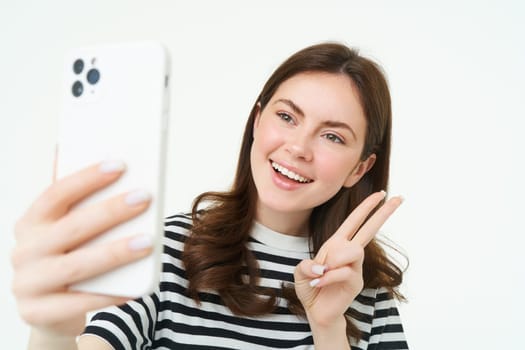 Portrait of young woman taking selfies on smartphone, posing for photo,. using mobile phone app for taking funny and cute pictures, isolated on white background.