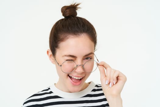 Close up portrait of stylish modern girl, wearing glasses, smiling and winking at camera, standing over white background.