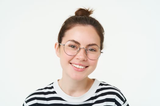 Close up portrait of cute young woman in glasses, looking at camera and smiling, trying on new spectacles, standing over white background.