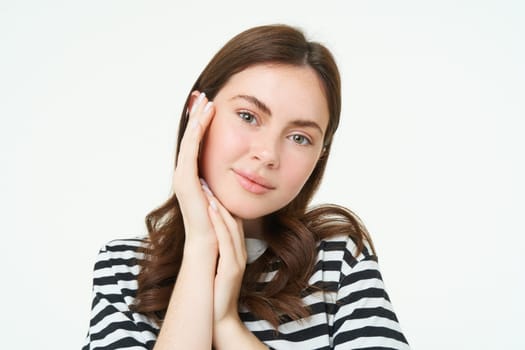 Portrait of tender, beautiful young woman, touches her clear, glowing skin, without blemishes or acne, concept of makeup and cosmetology, white background.