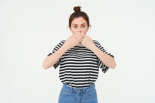 Image of shocked woman, covers her mouth, looks surprised, stands over white background. Copy space