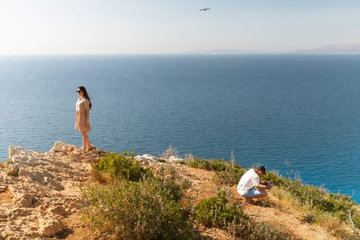 A young Caucasian guy films his happy girlfriend squatting in a dress and sunglasses, standing with her back to him, on a drone on the top of a mountain against the backdrop of a blurred sea, close-up side view.