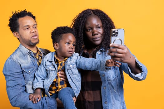 Happy african american parents take pictures with their young kid, posing together to capture sweet moments. Young proud mother and father take photos for memories on mobile phone.