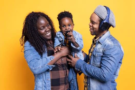 Happy mom and dad listening to their young kid sing a song in studio, african american father holding his microphone and wearing headset. Sweet family of three performing melodies.