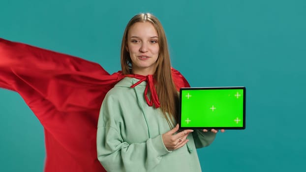 Joyous girl portraying superhero with red cape showcasing green screen tablet, studio backdrop. Joyful teenager posing as hero in costume on advertising chroma key digital device, camera A