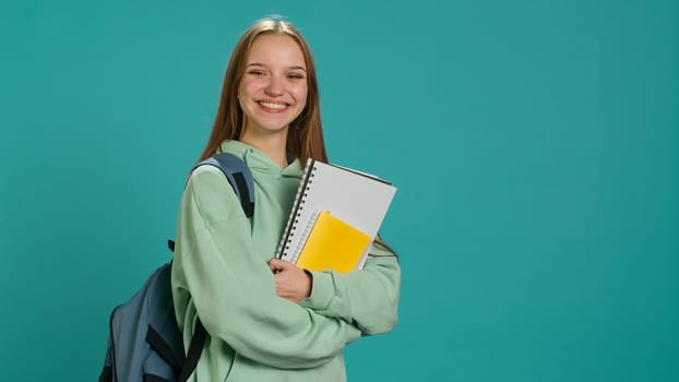 Portrait of radiant teenager with rucksack grinning, holding school notebook and notes, studio backdrop. Upbeat schoolgirl with textbook in arms, preparing to go to high school, camera A