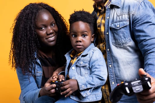 African american parents playing video games with their boy on console, using joystick to have fun against yellow background. Cheerful loving young family enjoying their leisure time.