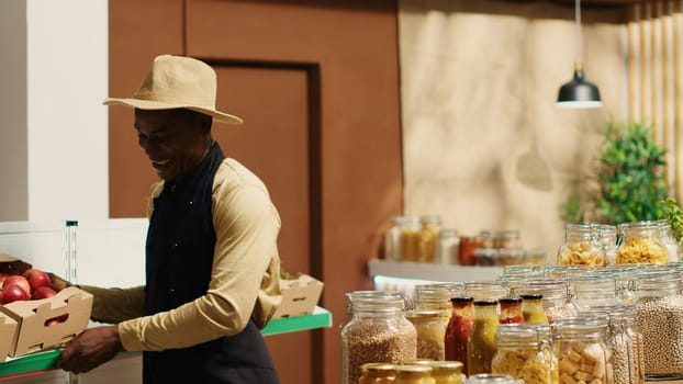 Business owner restocking crates of fresh fruits and vegetables, presenting organic locally grown produce to woman shopping at store. African american people chatting about vegan food. Camera 1.