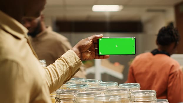 African american seller shows greenscreen display on mobile phone in local neighborhood market, selling natural bulk products. Merchant holds device with blank copyspace mockup screen. Camera 1.