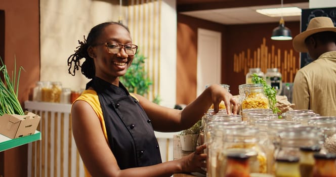 Local merchant organizing homemade pasta in reusable jars at zero waste supermarket, wearing apron and welcoming customers. African american woman in eco friendly store. Camera 2.