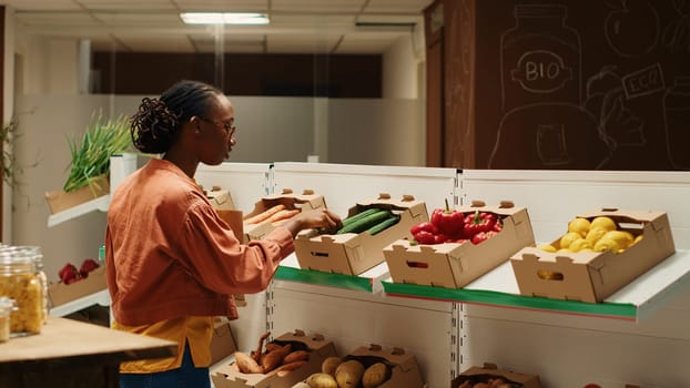 Vegan person searching for freshly harvested vegetables from crates, shopping for natural groceries at local zero waste eco store. African american client looking at organic food items. Camera 1.