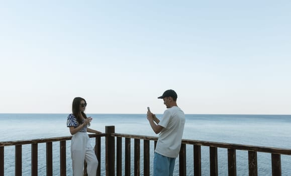 One cute guy in a cap takes a smartphone photo of a beautiful brunette girl in sunglasses standing on a wooden terrace against the backdrop of the sea on a sunny summer evening, close-up side view.