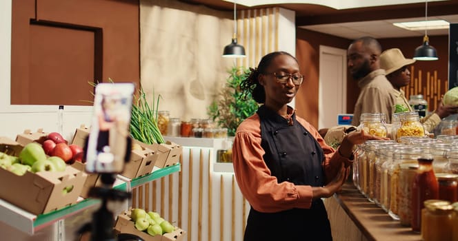 Merchant creating promotional video in local farmers market, talking about natural homemade food sauces and pantry supplies or dressings. African american woman working on marketing. Camera 2.