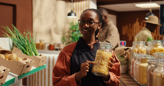 African american vendor presenting variety of pasta in jars, discussing about ethically sourced products and homemade food alternatives. Filming promotional video for healthy lifestyle. Camera 2.