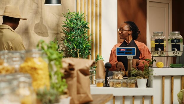 Merchant placing all produce on electronic scale at checkout, weighting fruits and veggies for regular client at zero waste eco store. African american people chat about healthy nutrition. Camera 1.