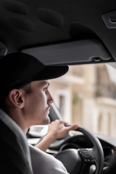 A beautiful view of one young Caucasian guy in a cap sitting half sideways behind the wheel of a car driving along the road on a summer day and looking to the left in the rearview mirror, close-up side view.