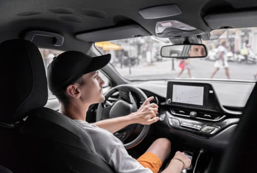 A beautiful view of one young Caucasian guy in a cap sitting half sideways behind the wheel of a car driving along the road on a summer day and looking to the left in the rearview mirror making a left turn, top side close-up view.