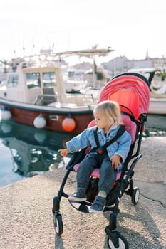 Little girl sits in a stroller on the shore and points to the sea. High quality photo