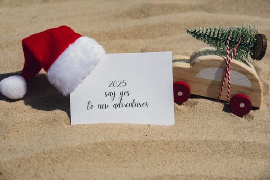 2025 SAY YES TO NEW ADVENTURES text on paper greeting card on background sandy beach sun coast. Christmas balls Santa hat New Year New Me Resolutions decoration. Summer vacation decor. Holiday concept calendar date postcard. Getting away Travel Business concept