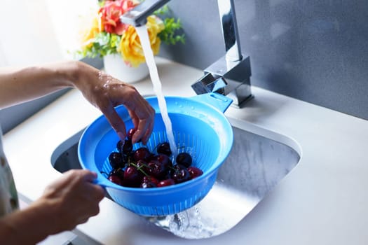 Unrecognizable woman washing ripe cherries in blue colander under water from faucet in kitchen sink