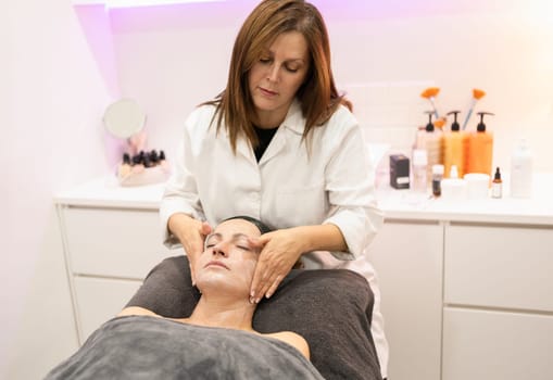 Young female beauty specialist massaging face with cosmetic cream of relaxed woman lying on table with closed eyes during skin care treatment in modern salon