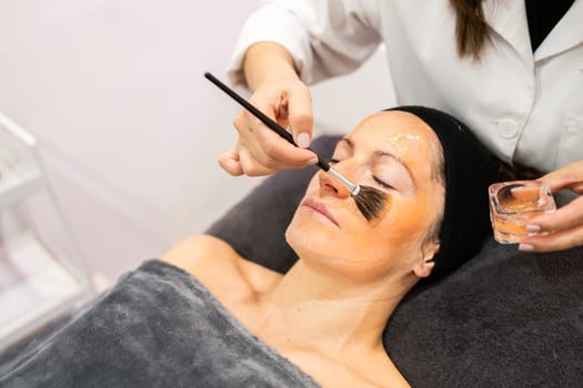 From above of crop professional cosmetologist applying moisturizing product on face of calm female client with brush in beauty center