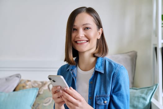 Young smiling woman using smartphone sitting on couch at home. Female looking at camera texting reading looking mobile applications technology internet online services for work study leisure lifestyle